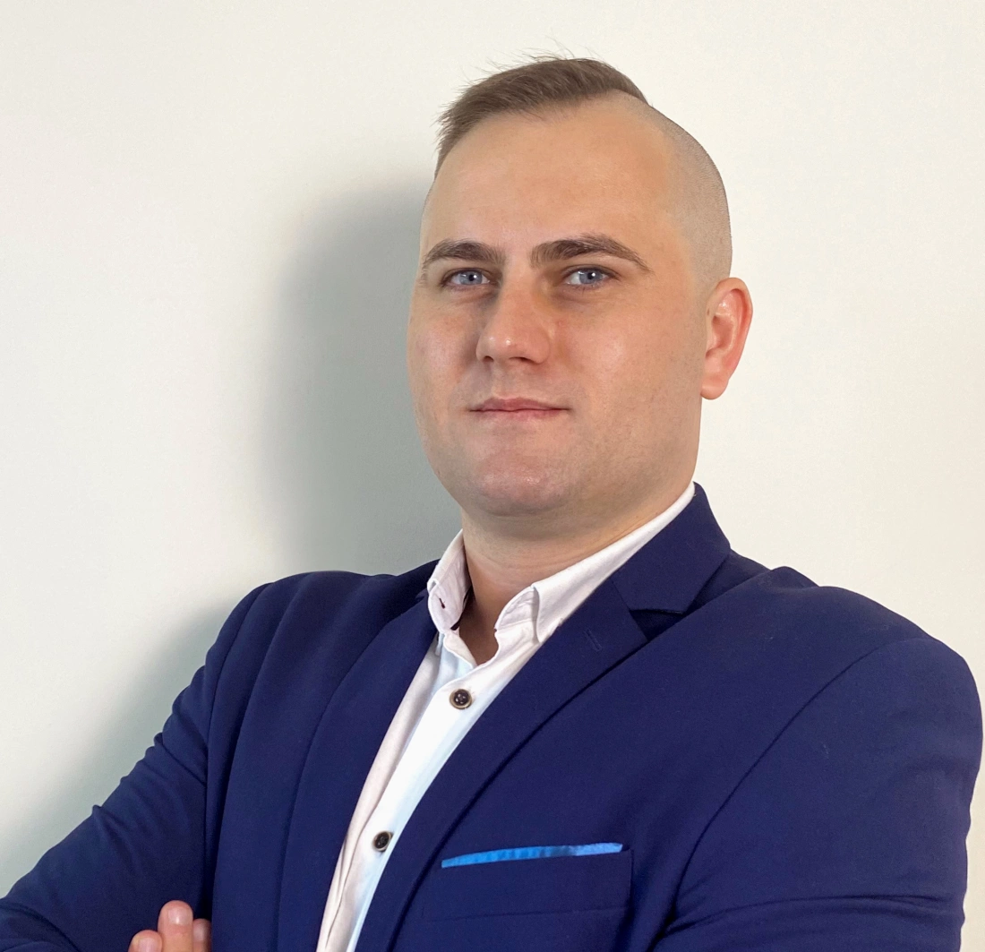 Mężczyzna w granatowym garniturze z kr&oacute;tko ostrzyżonymi włosami, stojący z założonymi rękami.