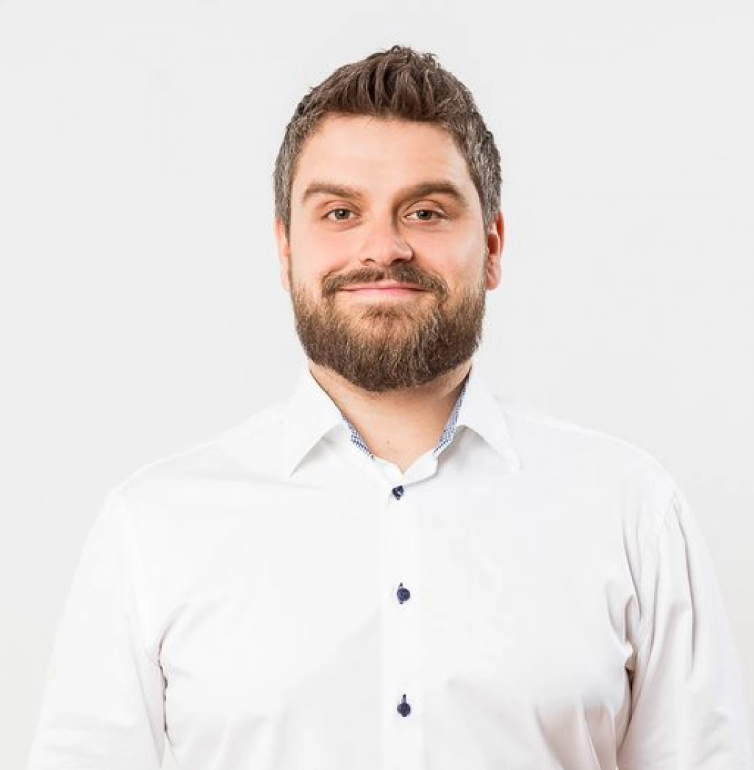 Mężczyzna z brodą w białej koszuli, uśmiechający się do kamery.