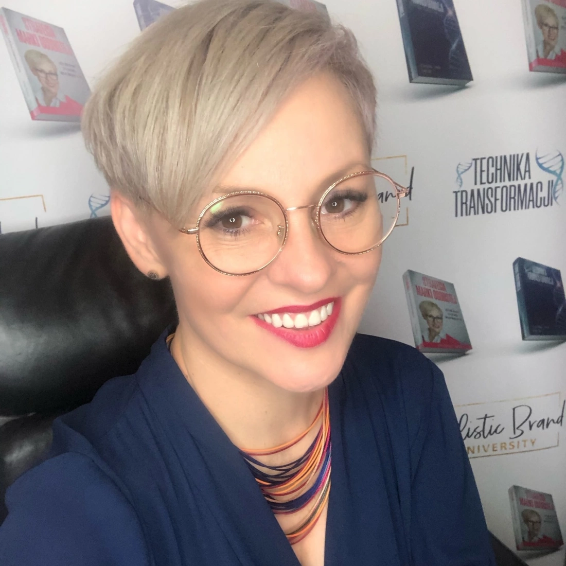 Kobieta w okularach, uśmiechająca się, z kr&oacute;tkimi, jasnymi włosami, siedzi przed tłem z książkami.