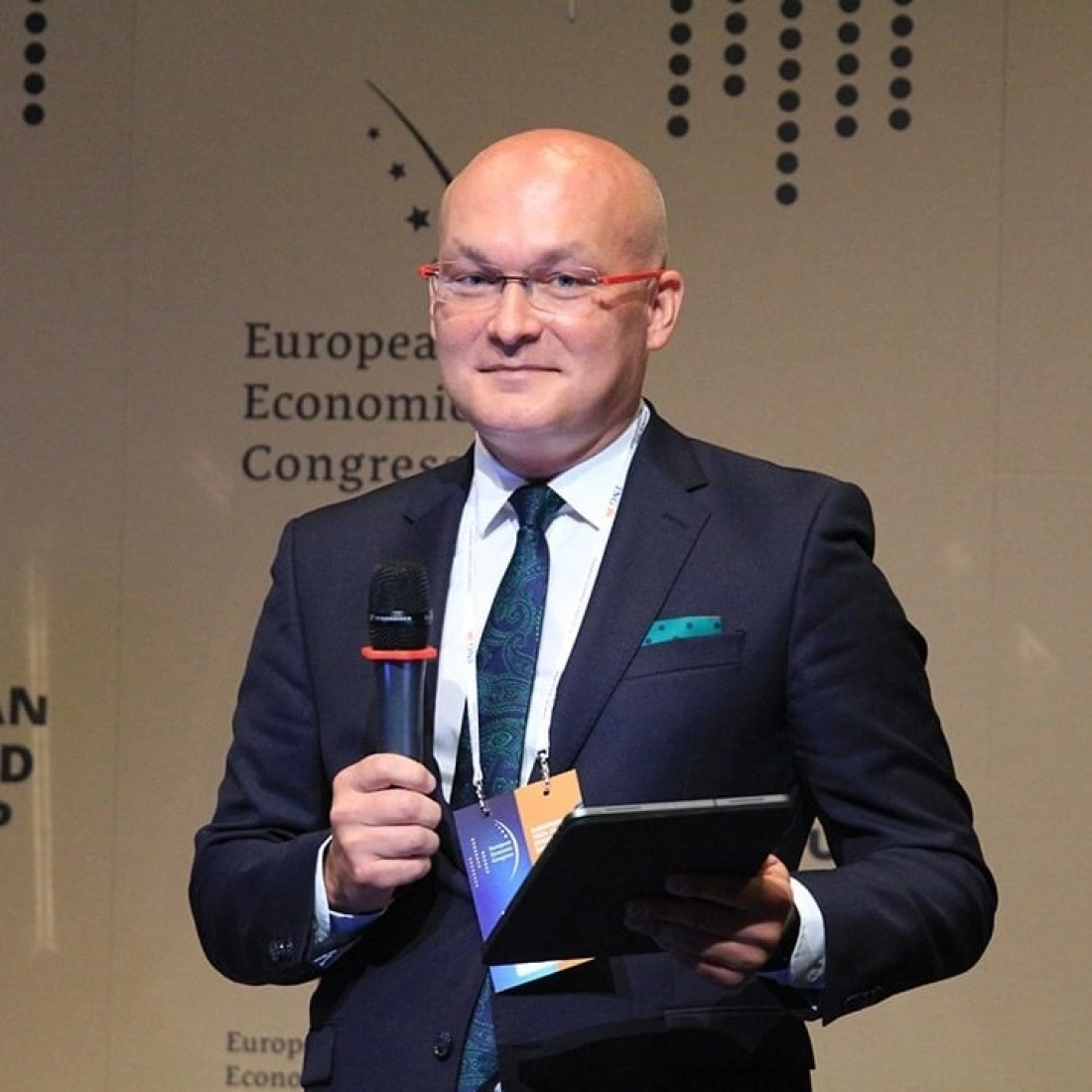 Mężczyzna w garniturze trzyma mikrofon i tablet, występując na tle napisu "European Economic Congress".
