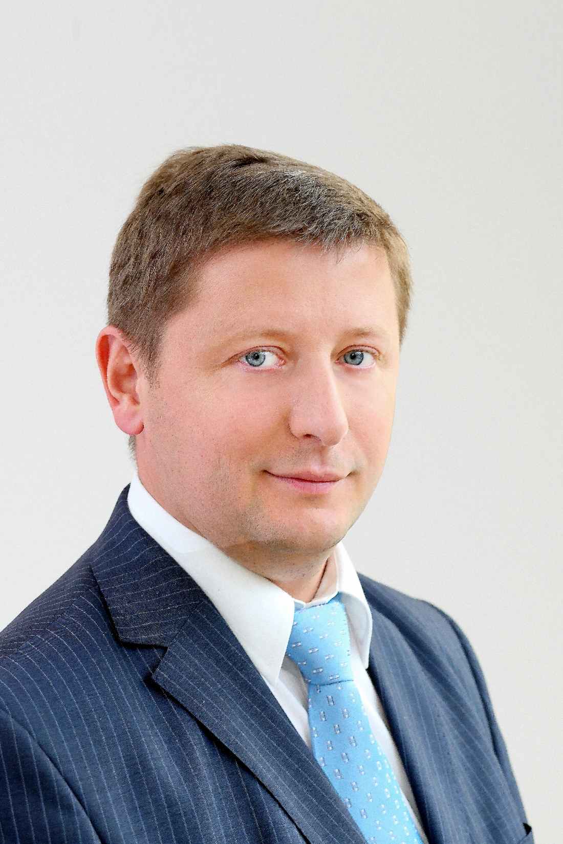 Mężczyzna w garniturze, z kr&oacute;tkimi włosami i niebieskim krawatem, patrzy w obiektyw. Tło jasne.