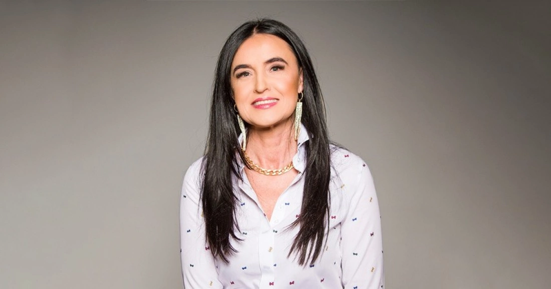 Kobieta uśmiechająca się na tle w jednolitym kolorze, ubrana w białą koszulę z wzorami.