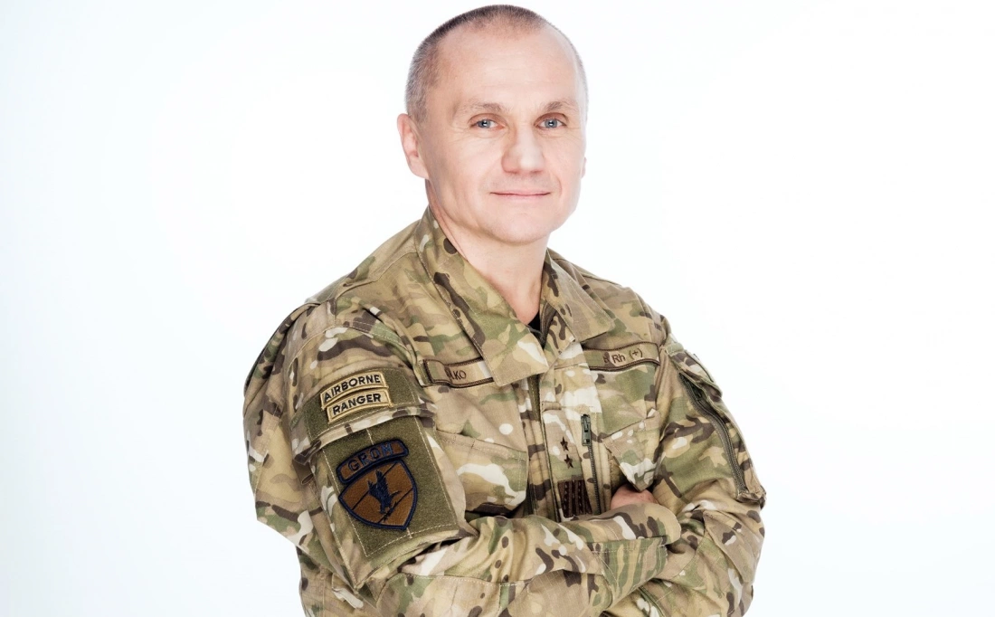 Mężczyzna w mundurze wojskowym z oznaczeniami spadochronowymi, uśmiechnięty, z założonymi rękami.