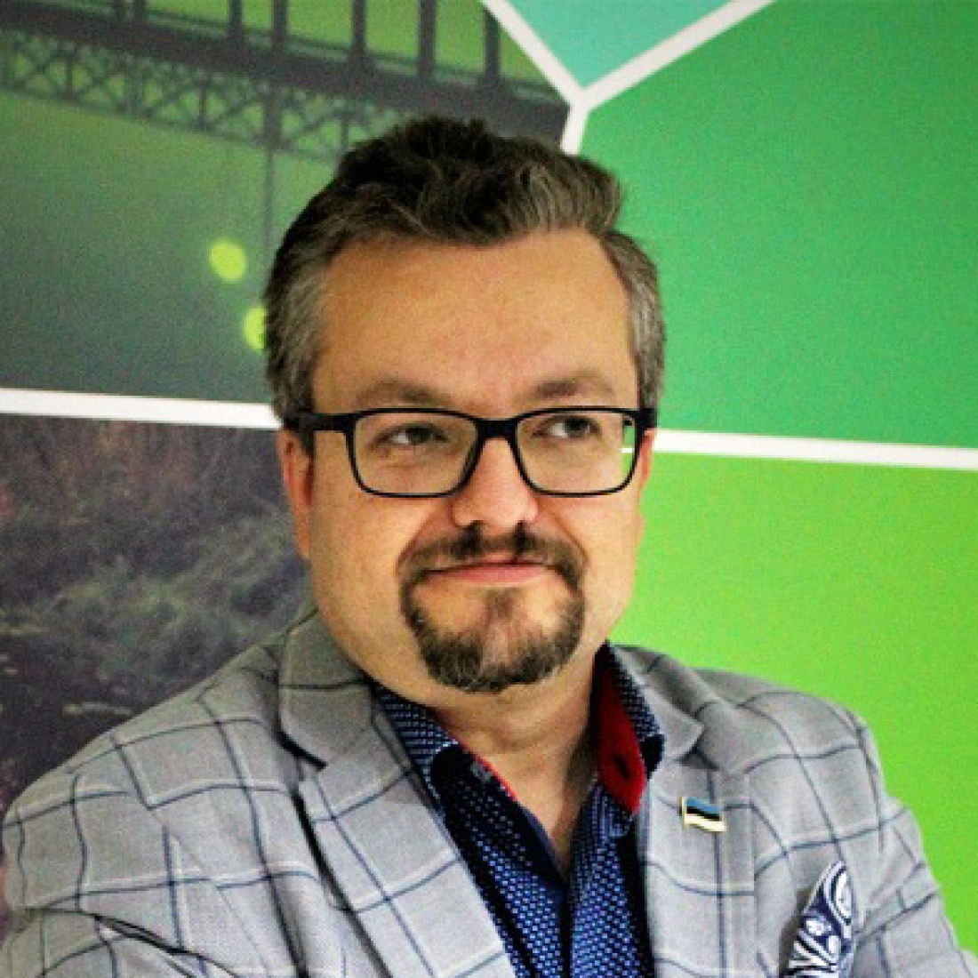 Mężczyzna w okularach z brodą, ubrany w elegancką marynarkę w kratę, stoi na tle kolorowej grafiki.