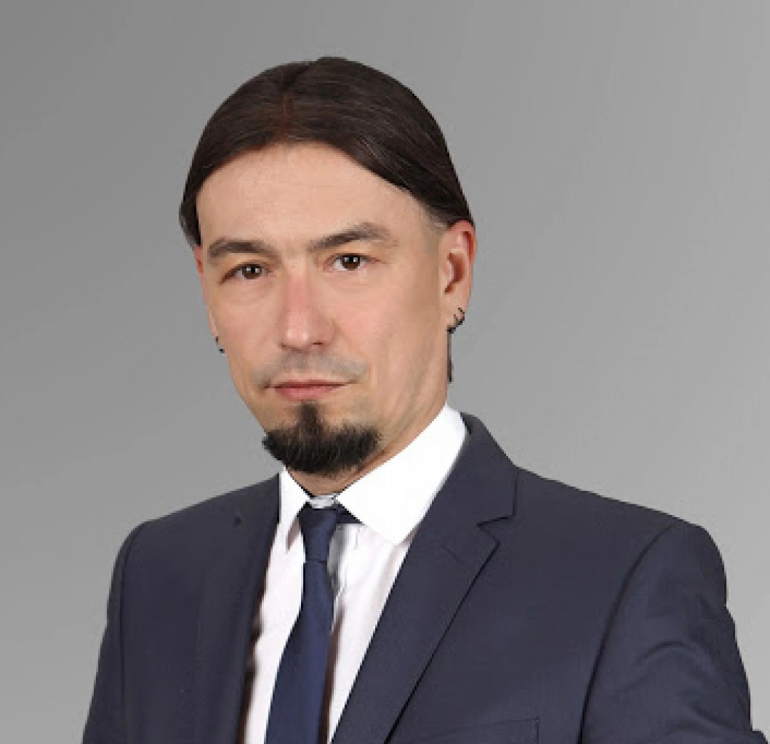 Mężczyzna w garniturze z długimi, ciemnymi włosami i zarostem, na jednolitym tle.