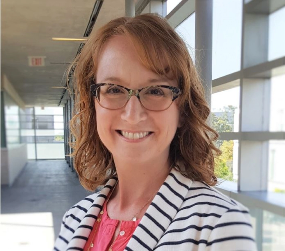 Kobieta w okularach z uśmiechem stoi w jasnym korytarzu.