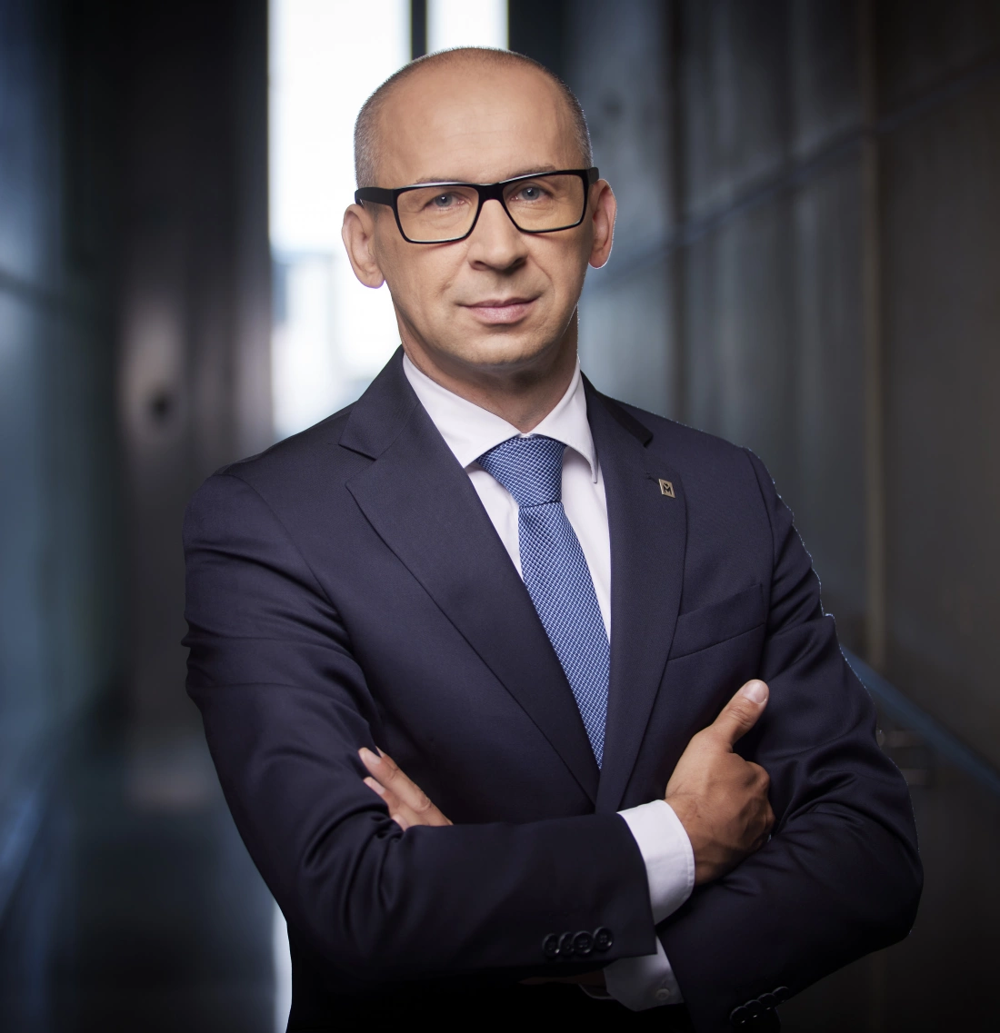 Mężczyzna w garniturze z założonymi rękami, stojący w nowoczesnym otoczeniu.