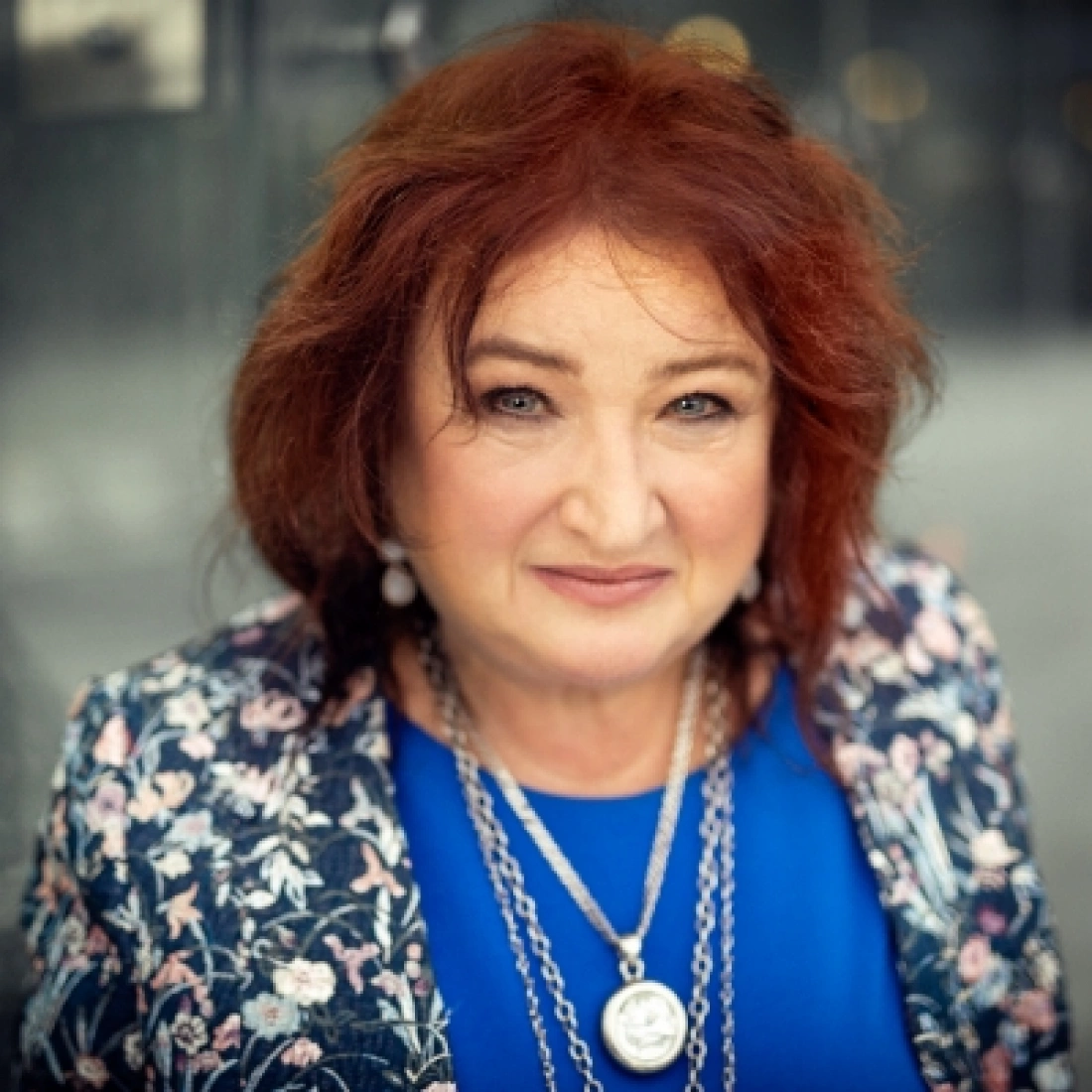 Uśmiechnięta kobieta z rudymi włosami, ubrana w niebieską bluzkę i ozdobną marynarkę.