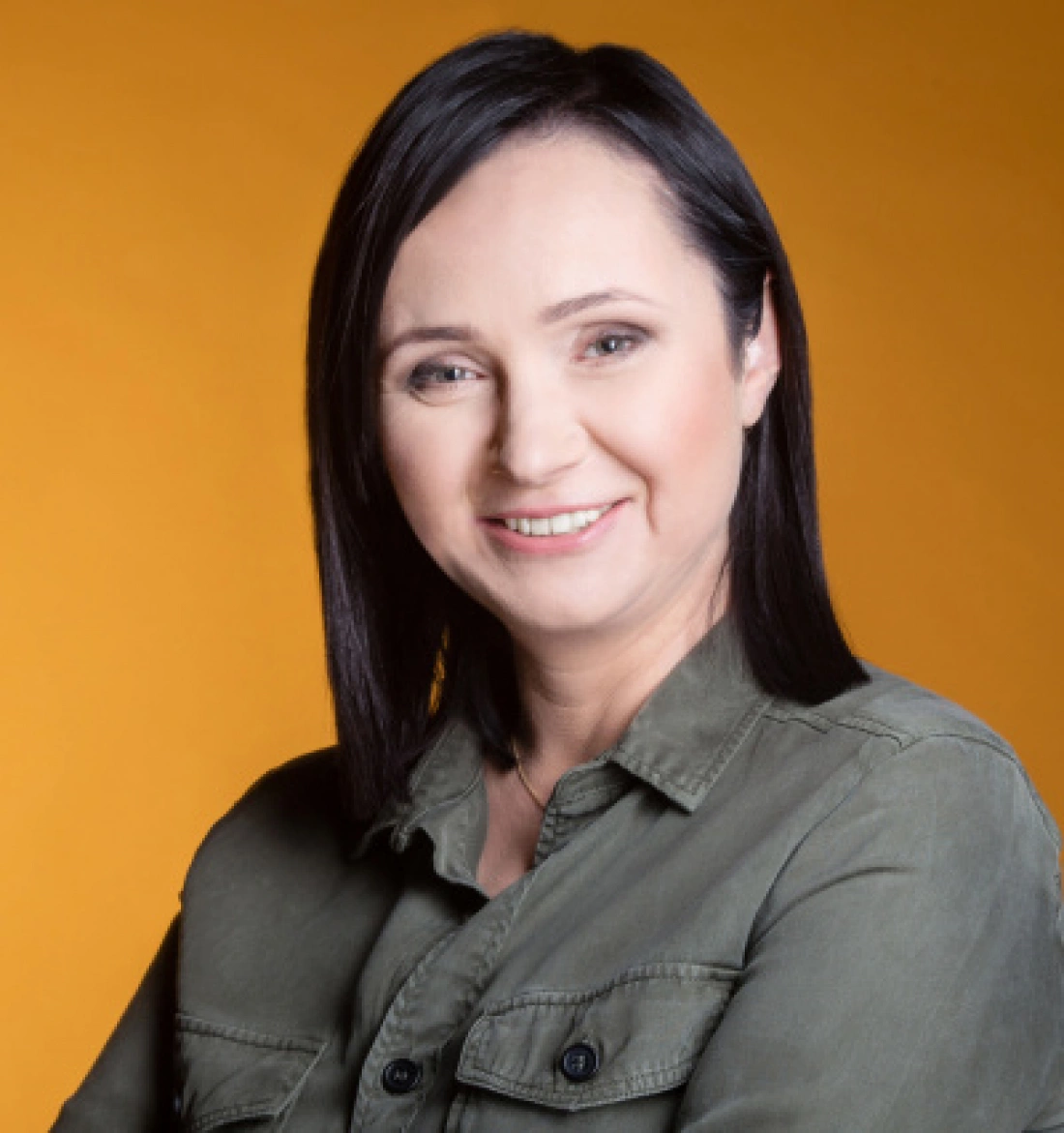 Kobieta z krótkimi, ciemnymi włosami w zielonej koszuli, uśmiechająca się na tle w kolorze pomarańczowym.