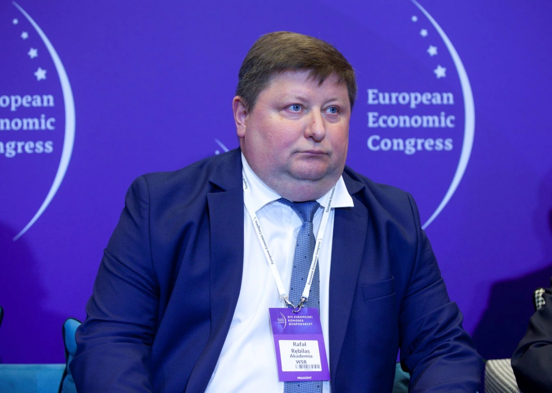 Mężczyzna w garniturze siedzi na tle fioletowego plakatu z napisami związanymi z Europejskim Kongresem Gospodarczym.