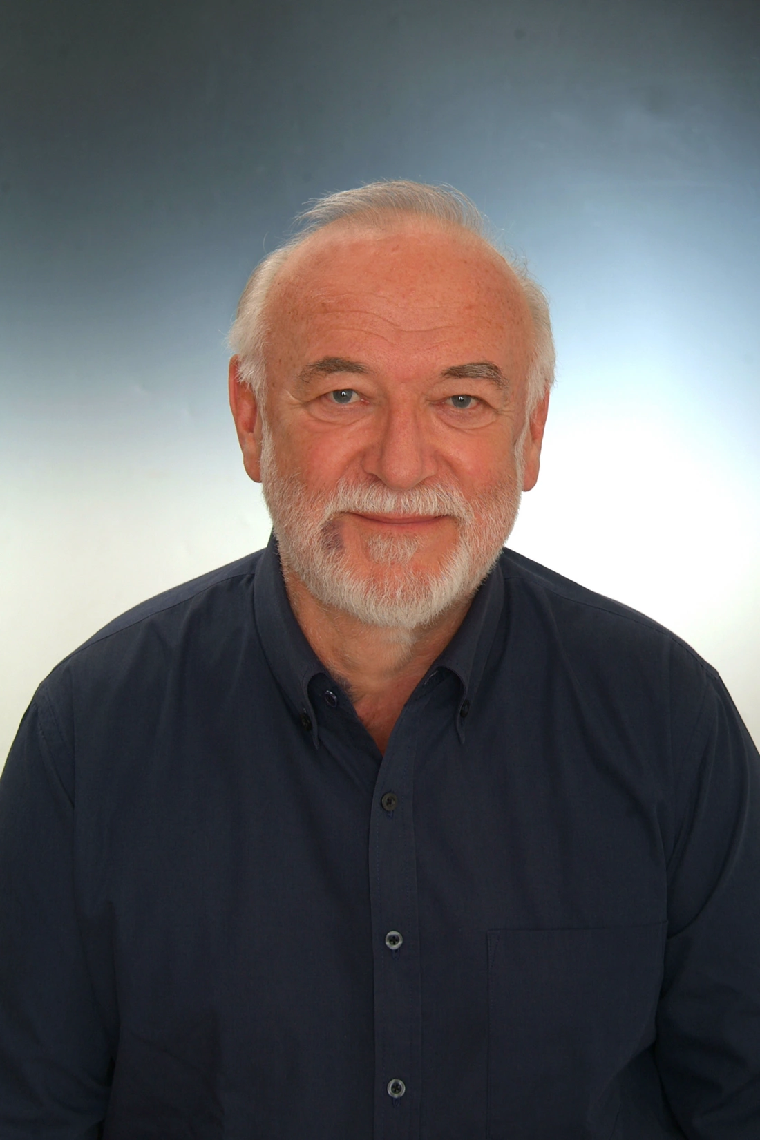 Mężczyzna z siwą brodą, uśmiechający się, w granatowej koszuli, na jasnym tle.