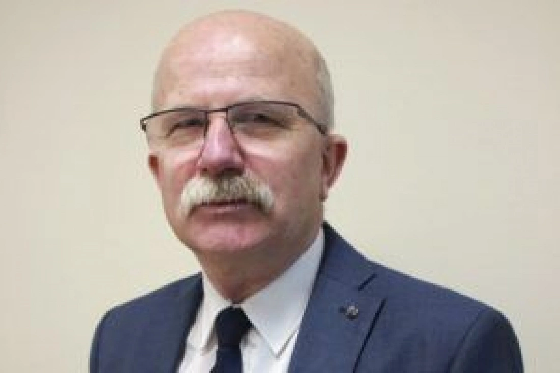 Mężczyzna w okularach, w garniturze, z siwą wąsami, stoi na neutralnym tle.