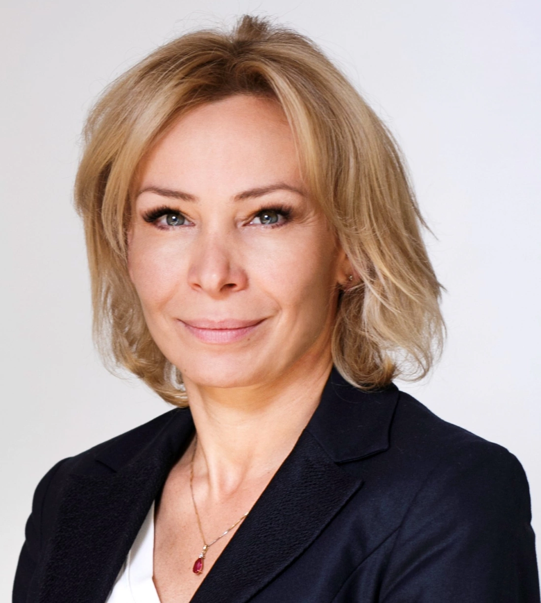 Kobieta o blond włosach w eleganckim ubraniu, uśmiechająca się na tle jasnego tła.