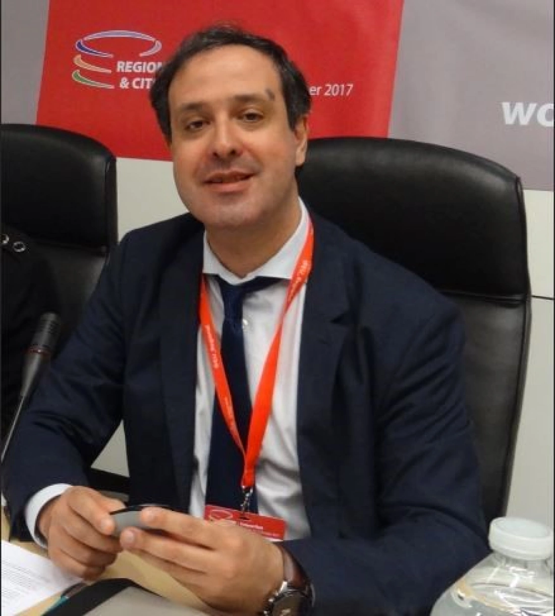 Mężczyzna w garniturze siedzący przy stole, uśmiechający się i trzymający telefon.