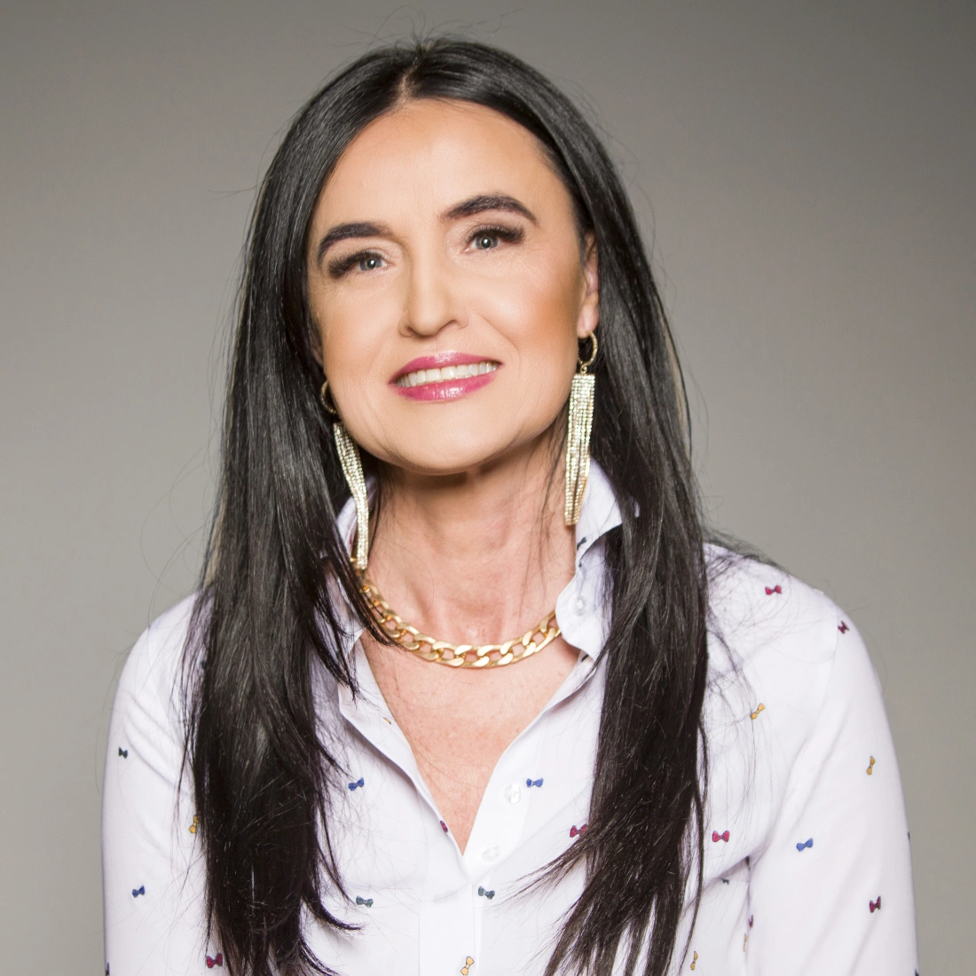 Kobieta z długimi, ciemnymi włosami, uśmiechająca się w białej koszuli z ozdobami i dużymi kolczykami.