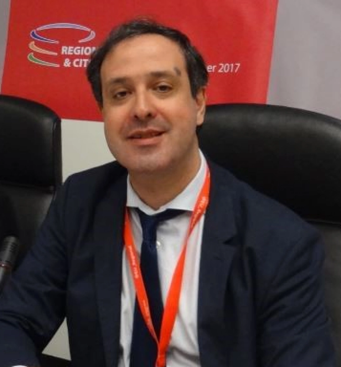 Mężczyzna w garniturze siedzący przy biurku, uśmiechający się do kamery.