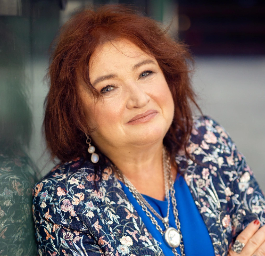 Kobieta z rudymi włosami, uśmiechająca się w eleganckim ubraniu z kwiatowym wzorem.