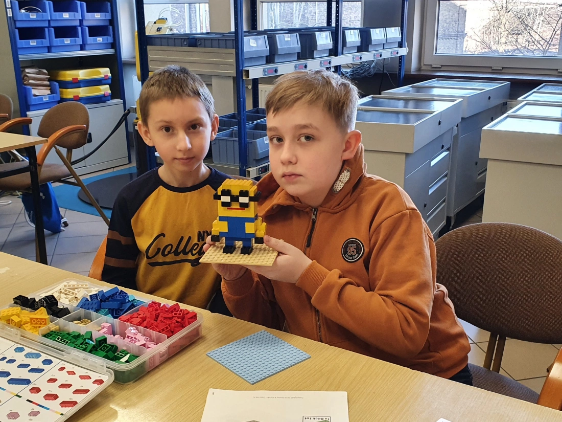 Dwaj chłopcy siedzą przy stole, trzymając model z klock&oacute;w LEGO, podczas zajęć w klasie. Na stole widoczne są r&oacute;żne klocki LEGO.