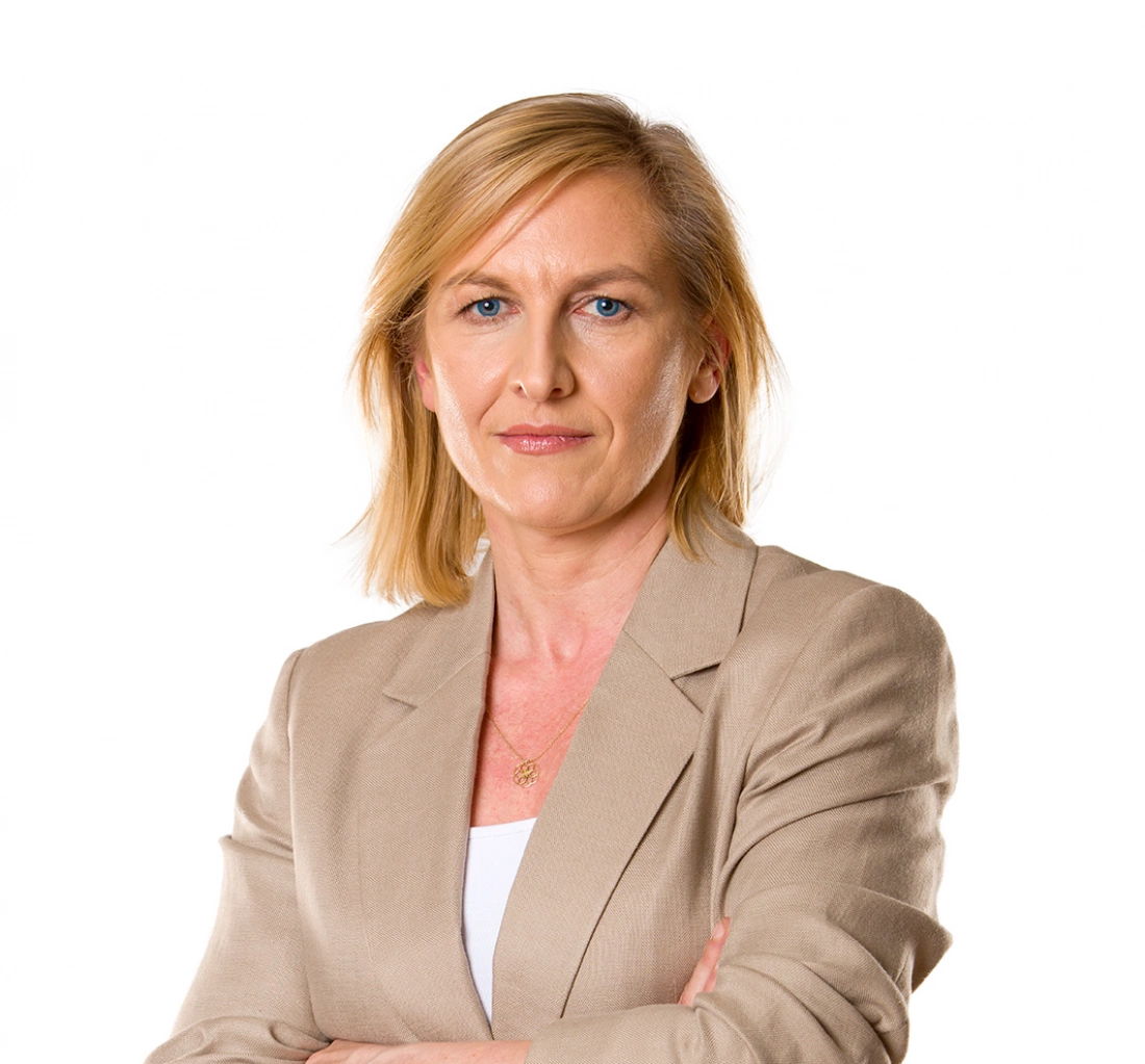 Kobieta w beżowym żakiecie z założonymi rękami, patrząca poważnie w kamerę.