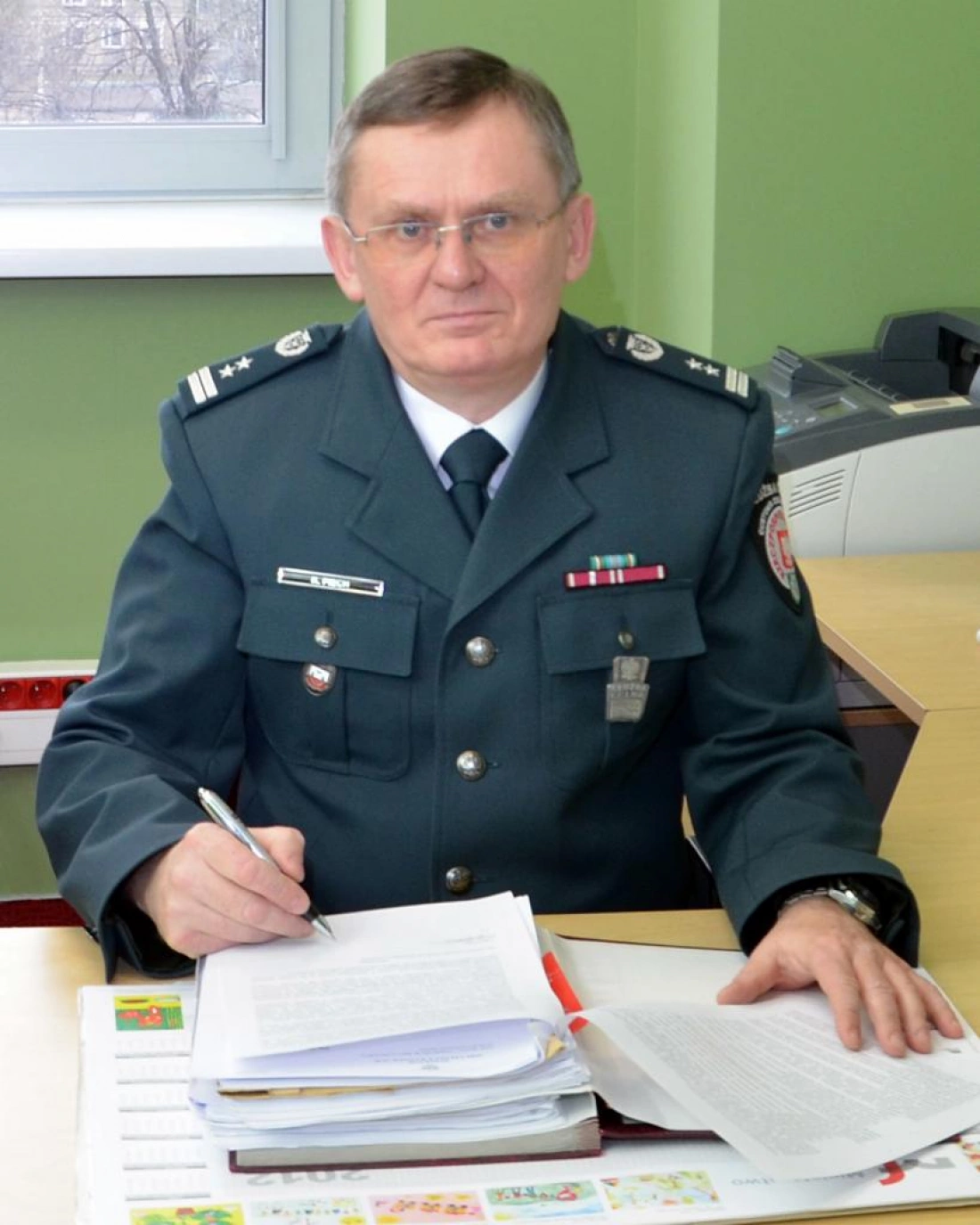 Mężczyzna w mundurze siedzi przy biurku, pisząc coś na dokumentach. W tle widać okno i elementy biurowe.