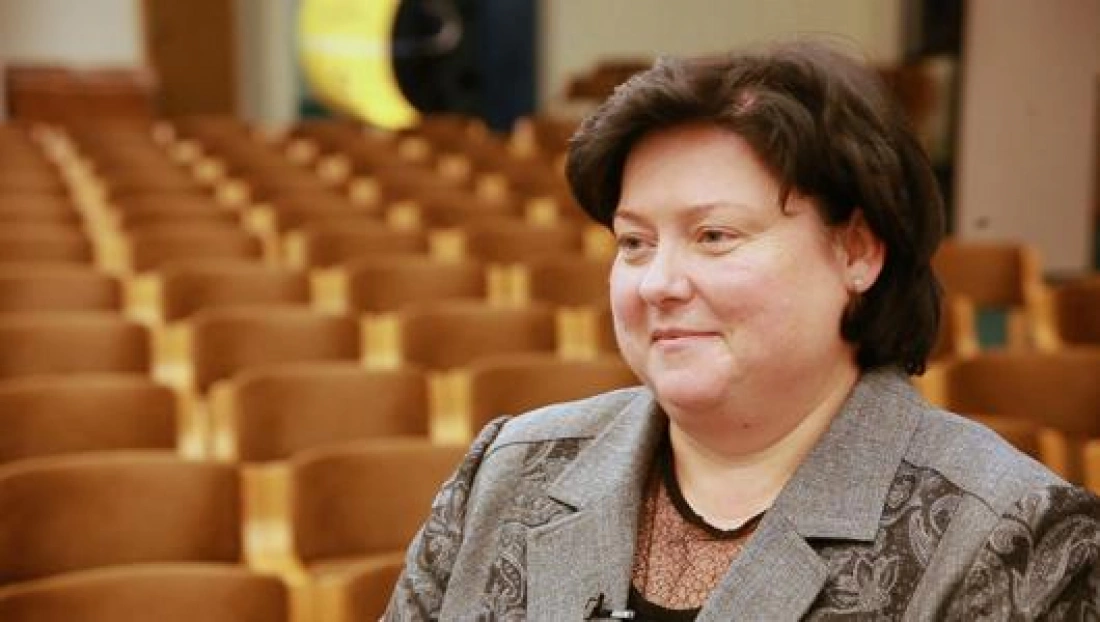 Kobieta w eleganckim stroju siedzi w sali z rzędem krzeseł w tle.