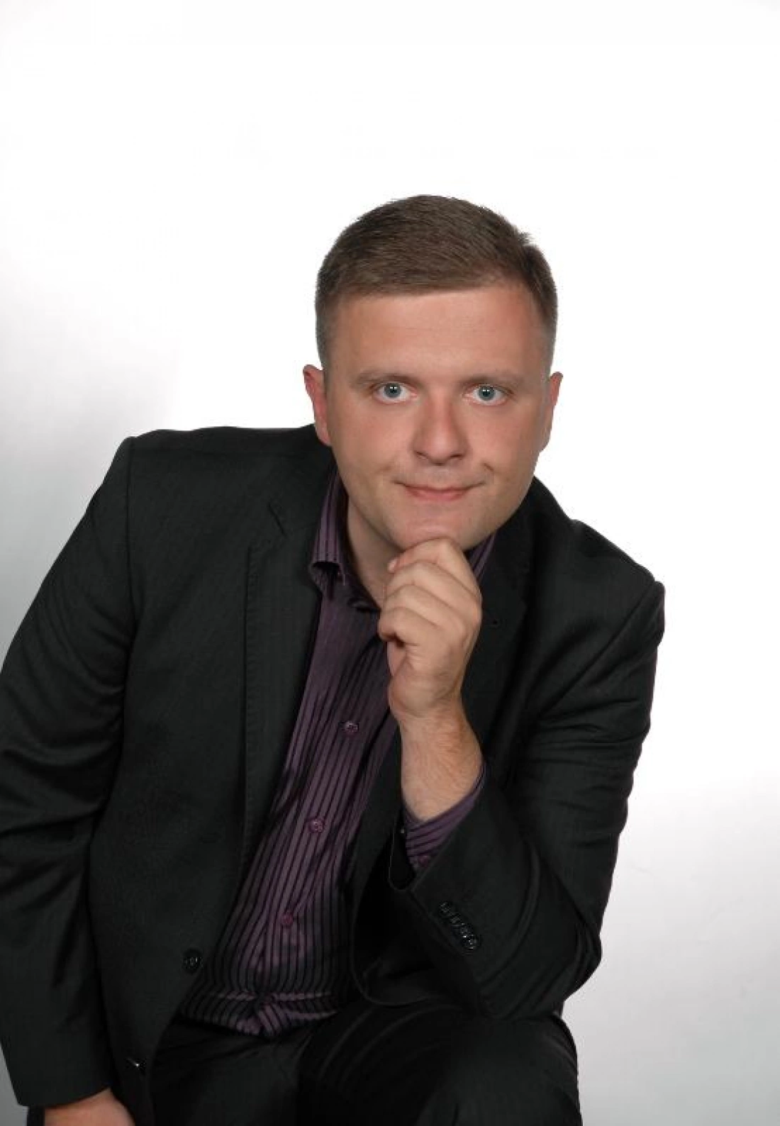 Mężczyzna w garniturze, siedzący z ręką przy brodzie, na tle białej ściany.