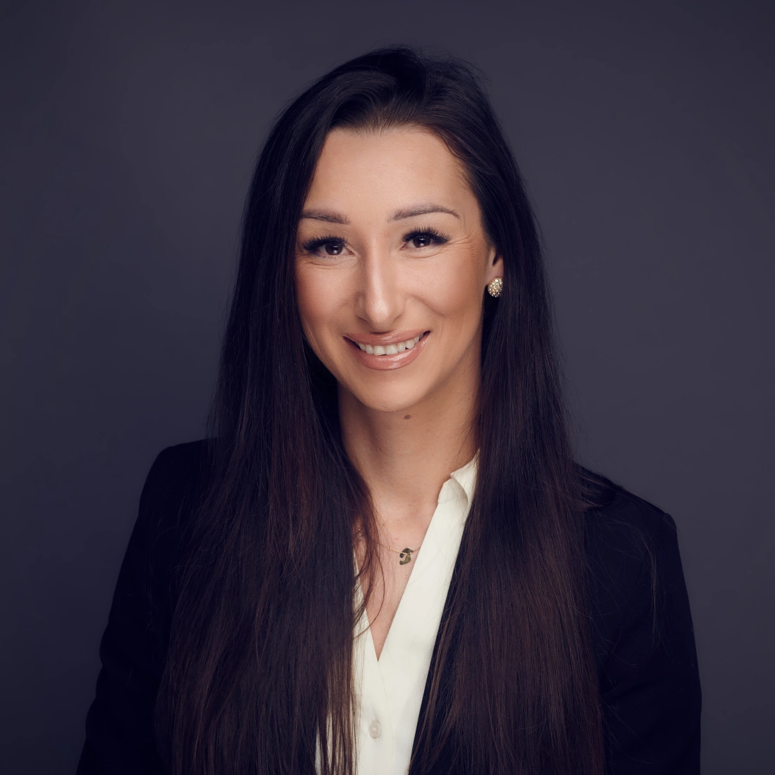 Portret kobiety w eleganckim ubraniu, uśmiechającej się na tle w ciemnym kolorze.