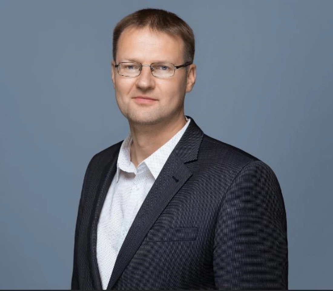 Mężczyzna w eleganckim garniturze, z okularami, stojący na jednolitym tle.