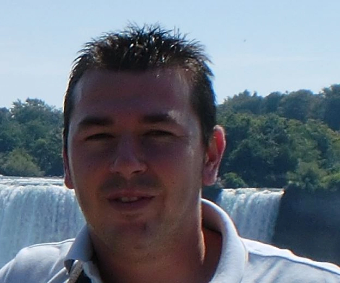 Mężczyzna w koszulce z krótkim rękawem, stojący przed wodospadem w słoneczny dzień.