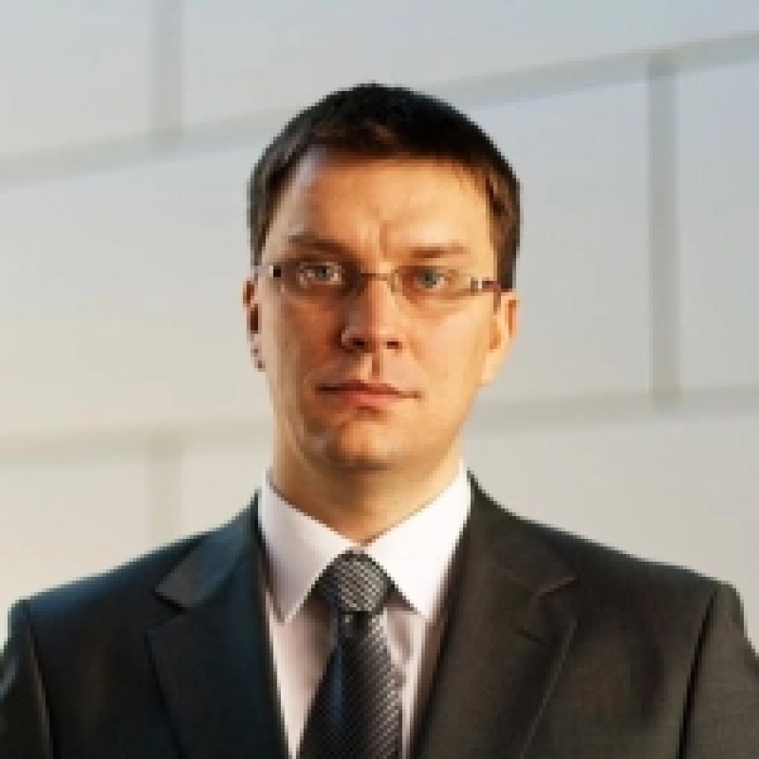 Mężczyzna w garniturze z okularami, stojący w oficjalnej scenerii.