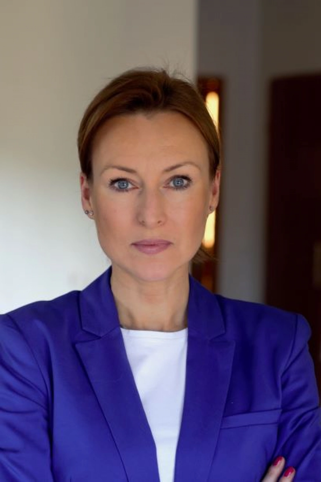 Kobieta w eleganckim niebieskim żakiecie, z poważnym wyrazem twarzy, stoi na tle oświetlonego wnętrza.
