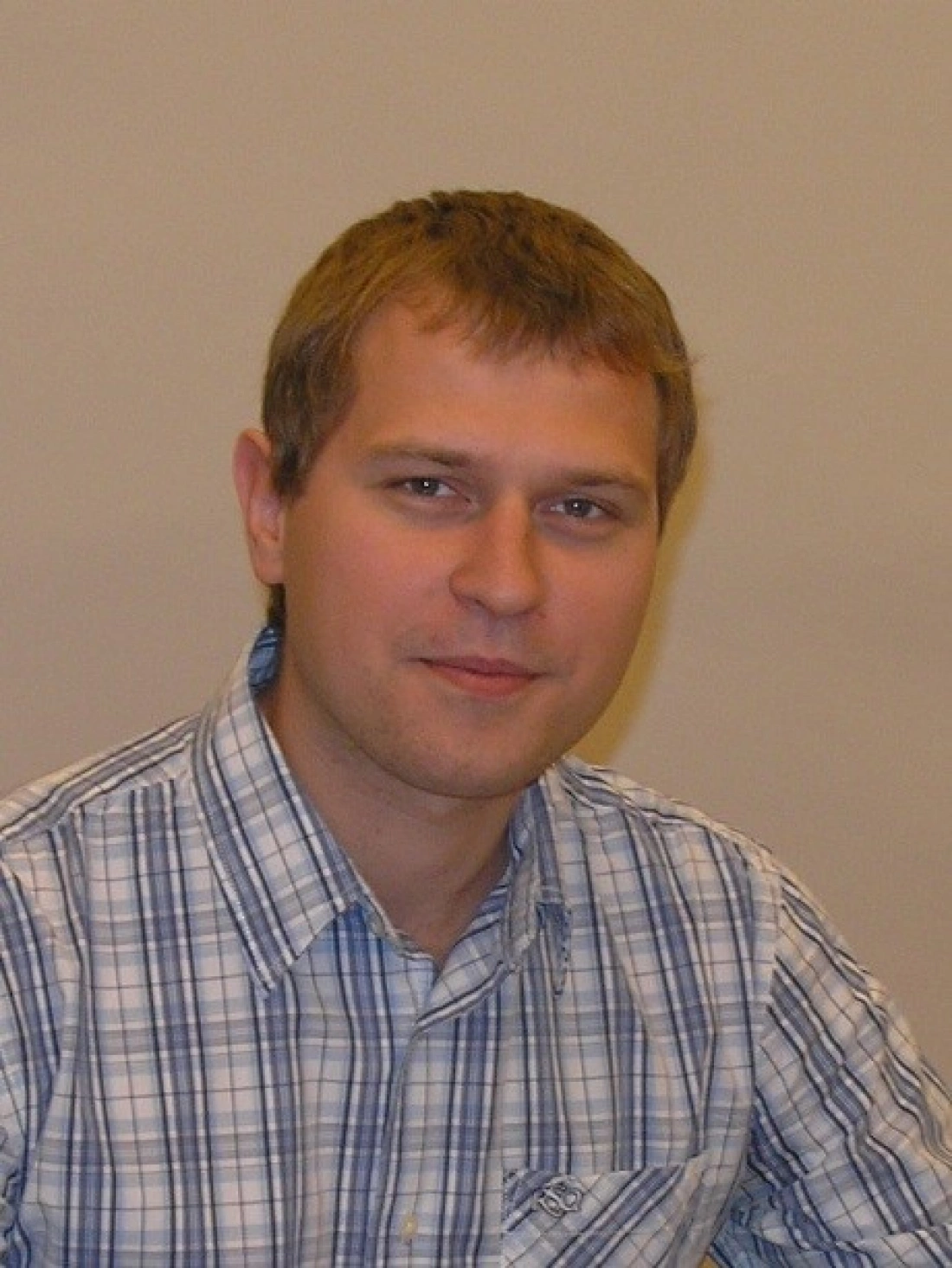 Mężczyzna w kratkowanej koszuli siedzi przy biurku, uśmiechając się do kamery.