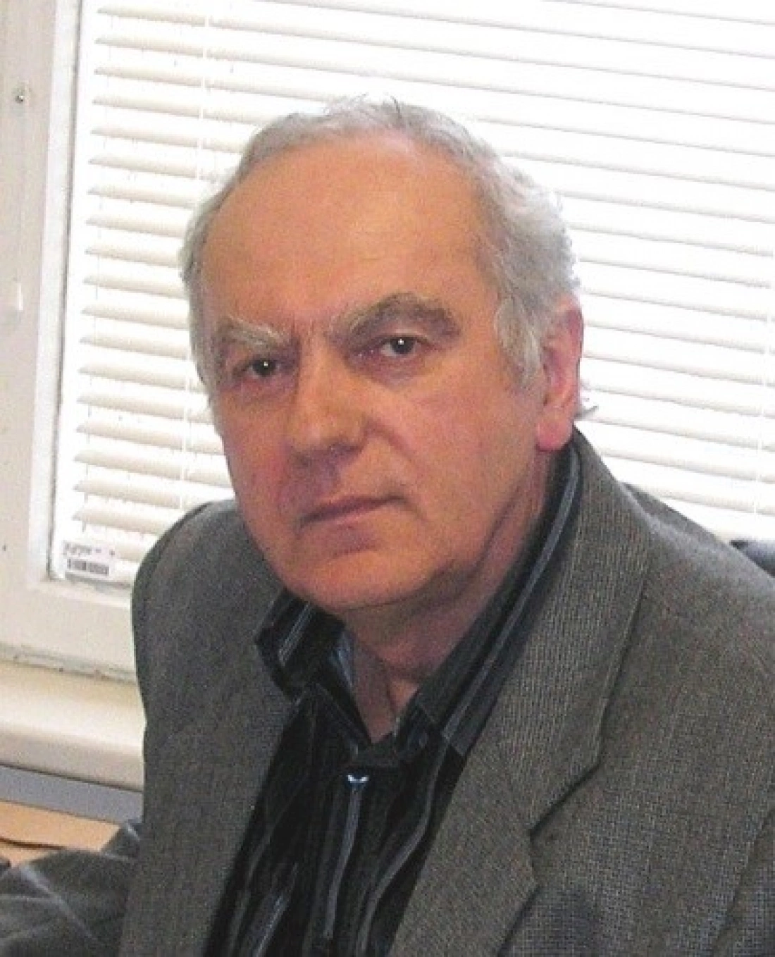 Mężczyzna w szarym garniturze, siedzący przy biurku, z poważnym wyrazem twarzy.