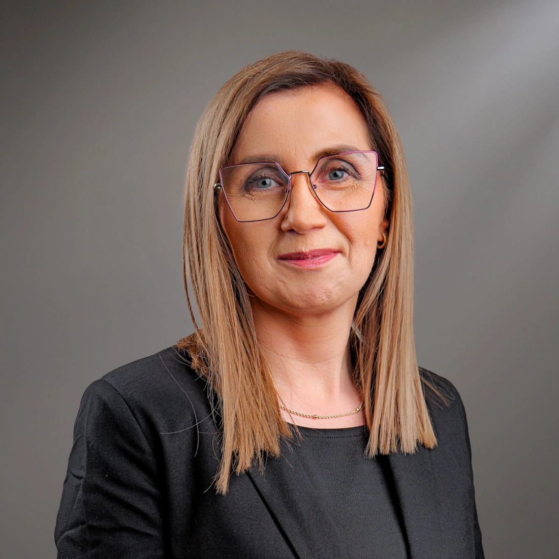 Kobieta z krótkimi, jasnymi włosami i okularami, uśmiechająca się na neutralnym tle.