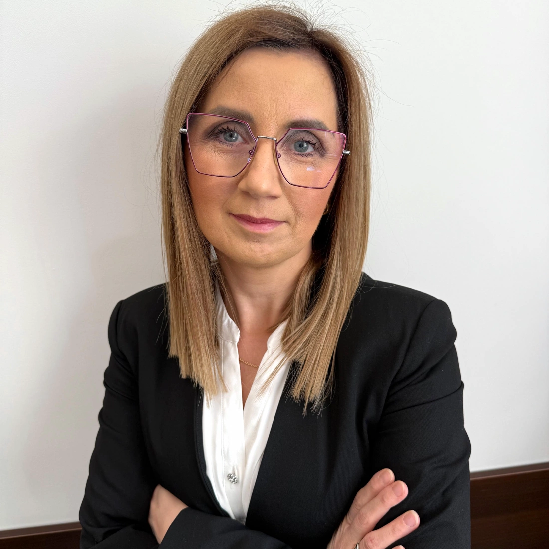 Kobieta z krótkimi, jasnymi włosami i okularami, uśmiechająca się na neutralnym tle.