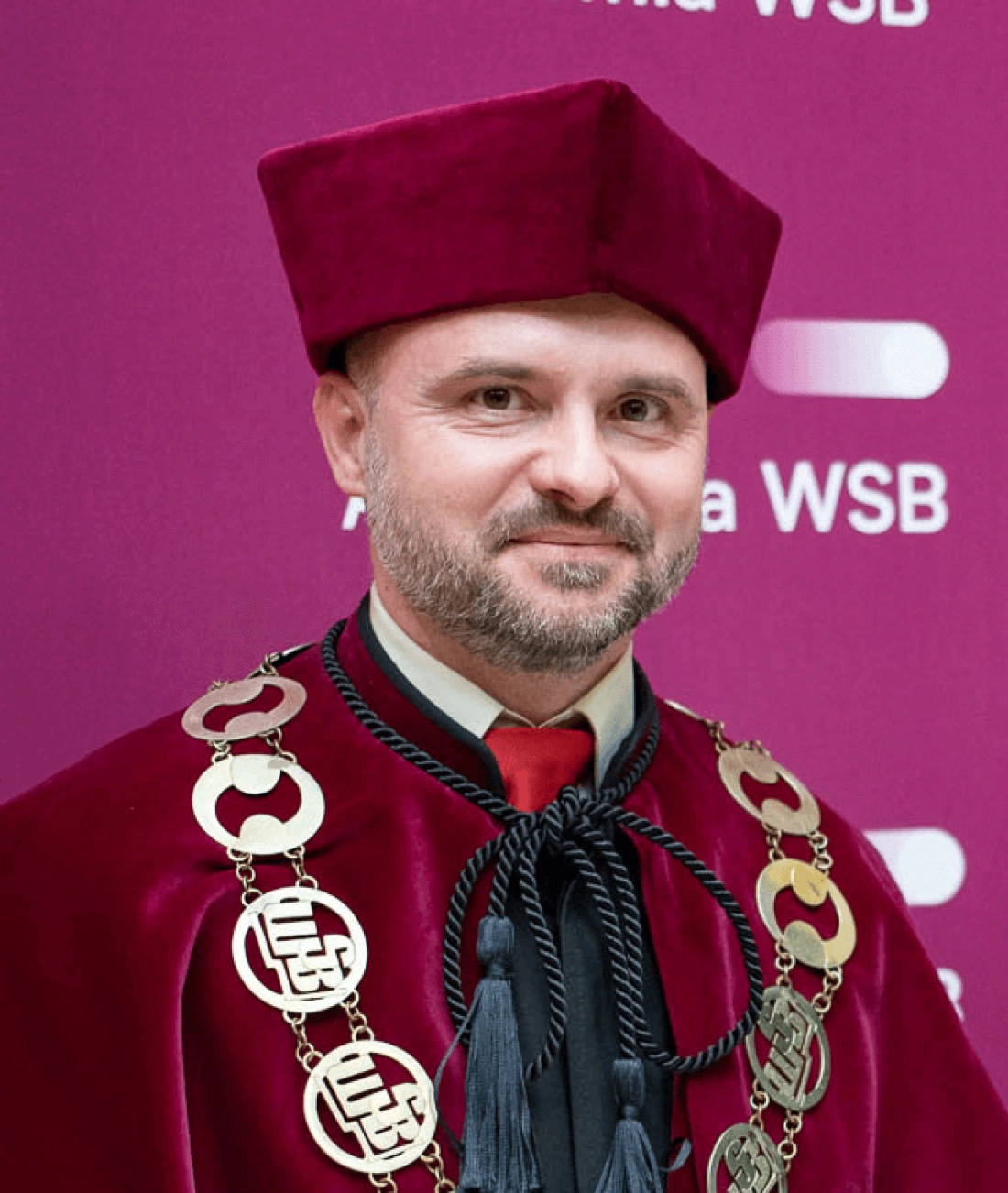 Mężczyzna w czerwonej todze i birecie, z medalionami na szyi, uśmiechający się przed różowym tłem.