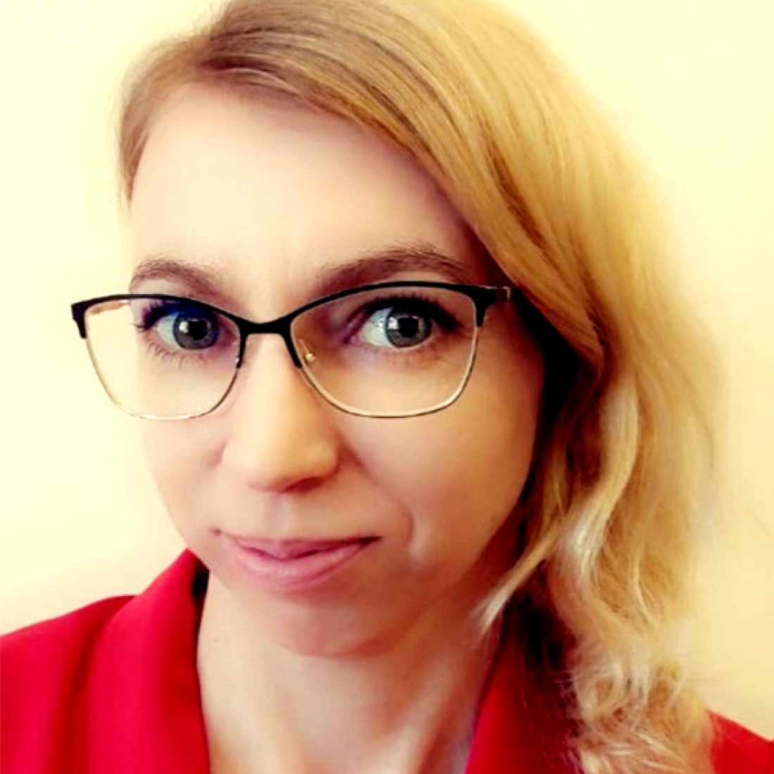 Kobieta z blond włosami w okularach, uśmiechająca się lekko, nosząca czerwoną bluzkę.