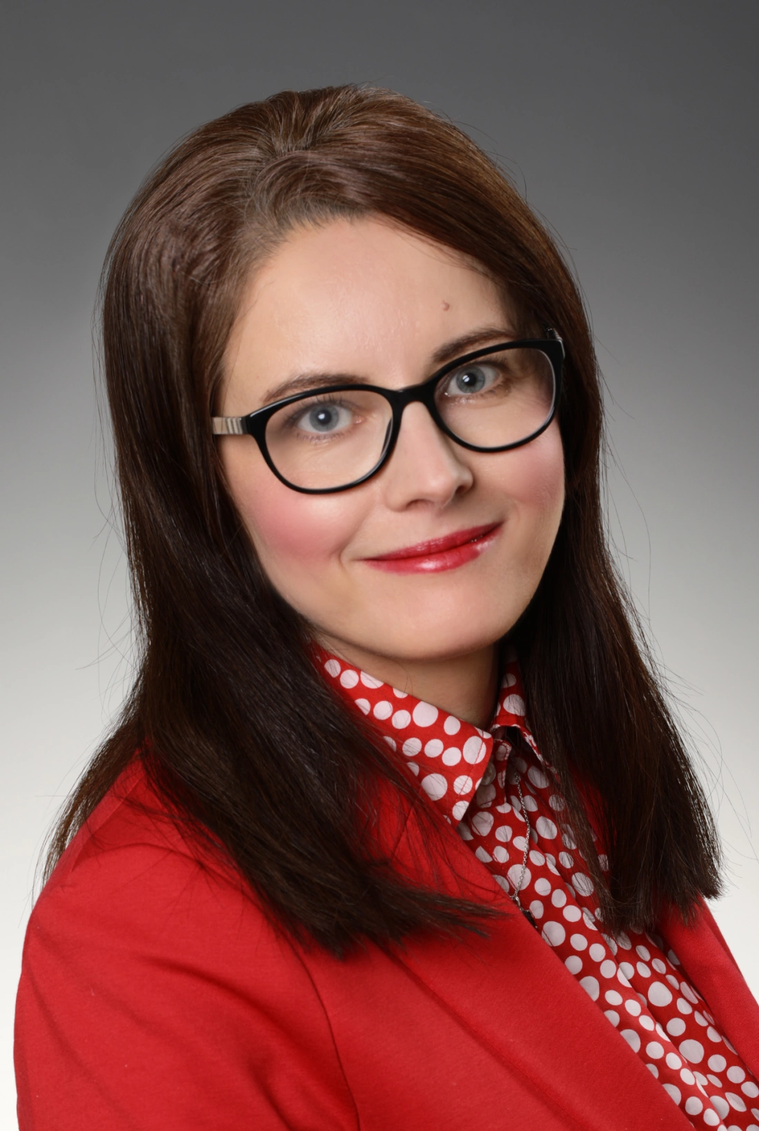 Kobieta w okularach z długimi włosami, uśmiechająca się, w czerwonej marynarce i koszuli w kropki.