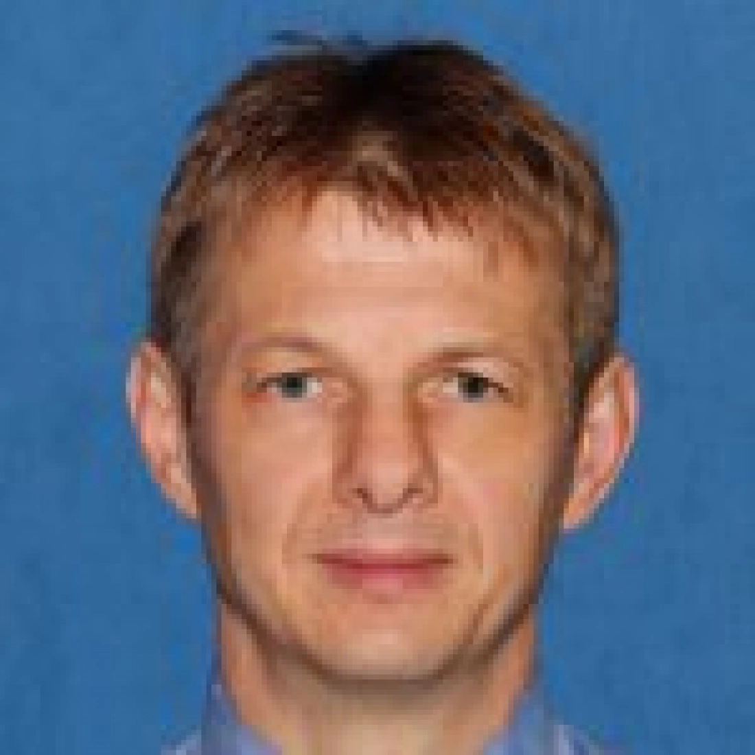 Mężczyzna z krótkimi, jasnymi włosami na tle niebieskiego tła.