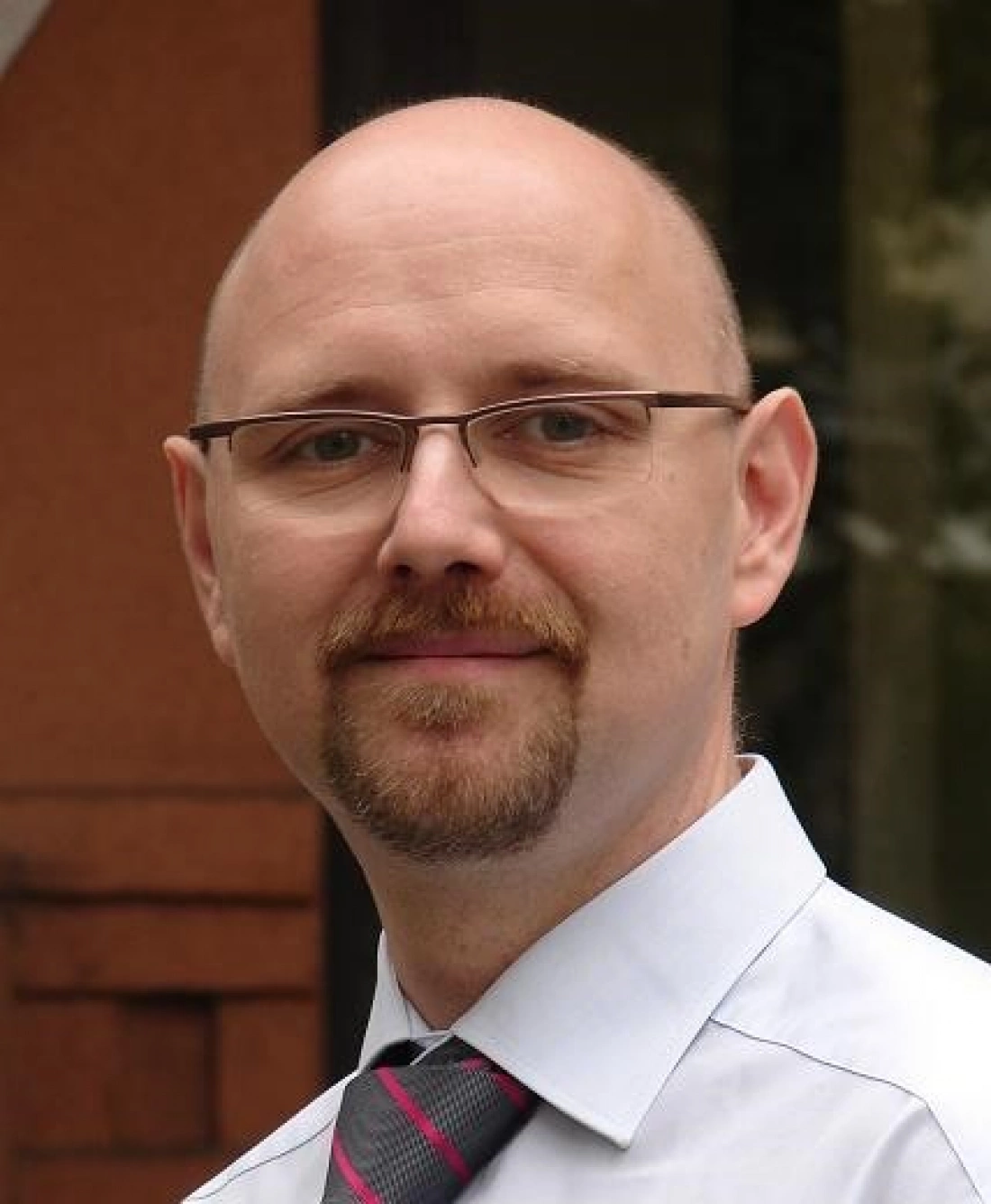 Mężczyzna z okularami, uśmiechający się na tle budynku.