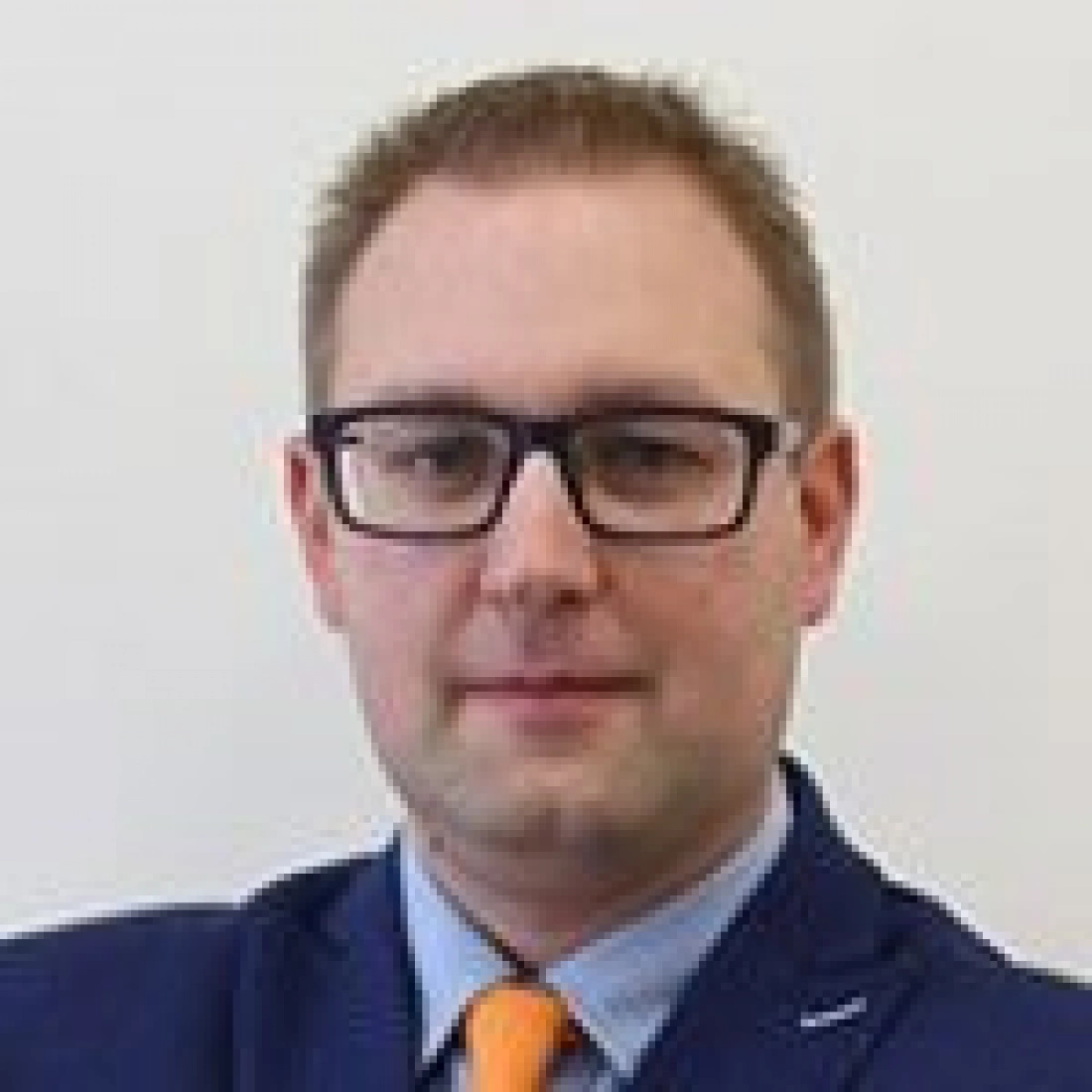 Mężczyzna w okularach, ubrany w granatowy garnitur i pomarańczowy krawat.