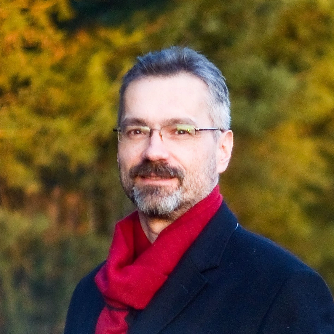 Mężczyzna z siwymi włosami i okularami, ubrany w czarny płaszcz i czerwoną chustę, uśmiecha się na tle zieleni.