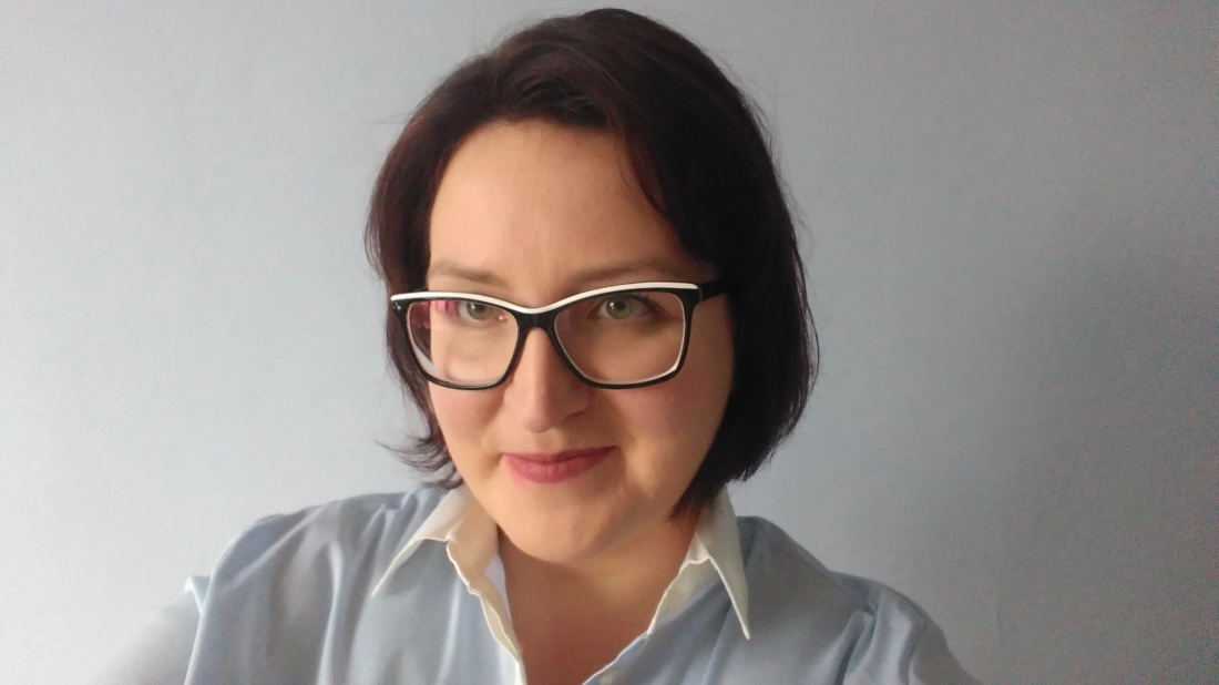 Kobieta w okularach z krótkimi, ciemnymi włosami, uśmiechająca się w jasnym pomieszczeniu.