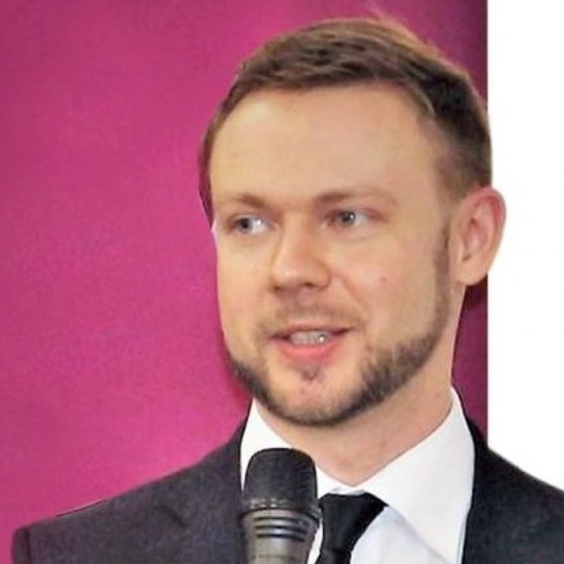 Mężczyzna w garniturze trzyma mikrofon, uśmiechając się na tle purpurowej ściany.