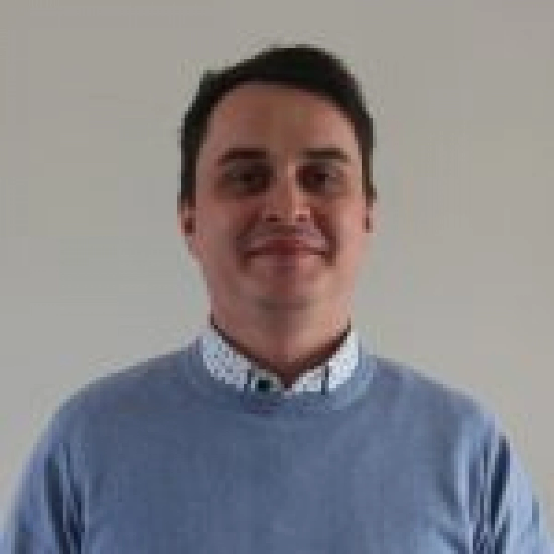 Mężczyzna w błękitnym swetrze z uśmiechem na tle jasnej ściany.