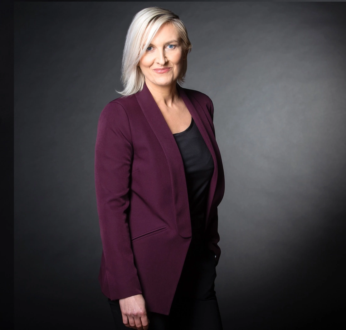 Kobieta w fioletowej marynarce, stojąca z uśmiechem na tle ciemnego tła.