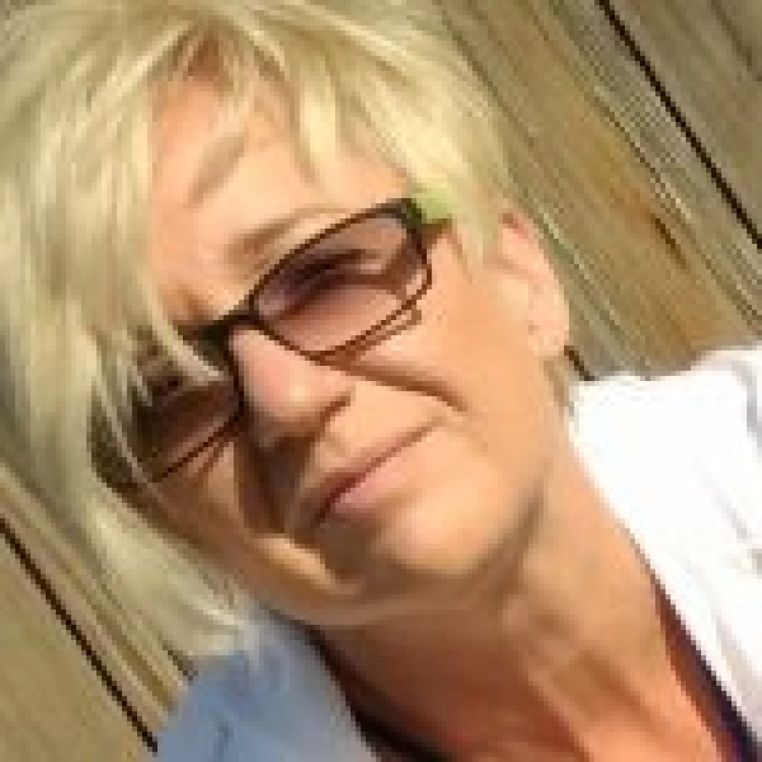 Kobieta z krótkimi, jasnymi włosami, w okularach, uśmiecha się do kamery.