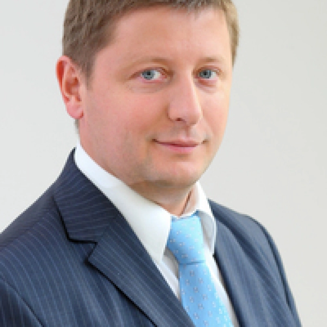 Mężczyzna w garniturze z niebieskim krawatem, uśmiechający się na tle jasnej ściany.