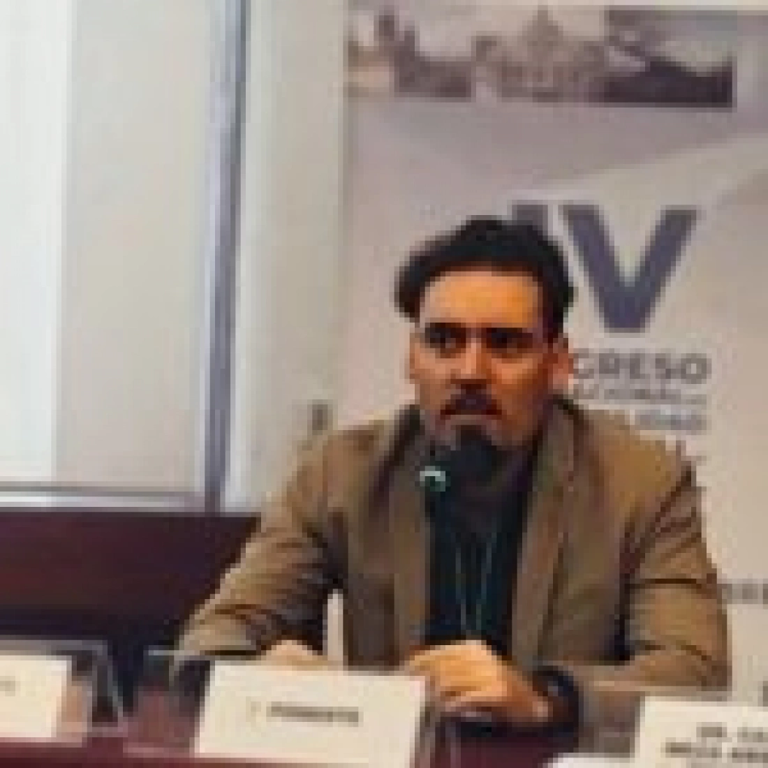 Mężczyzna siedzący przy stole, przemawiający do mikrofonu podczas konferencji.