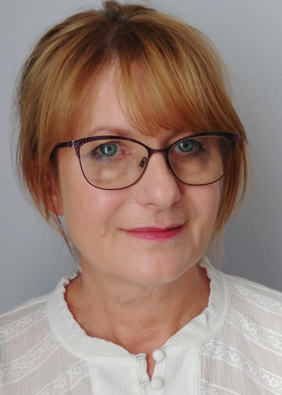 Kobieta z krótkimi blond włosami, w okularach, uśmiechająca się, na tle szarym. Ubrana w białą bluzkę.